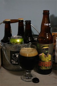 Glass of Dirty Commie Heathen along side the bottle with bottles and cans of other beers in the background
